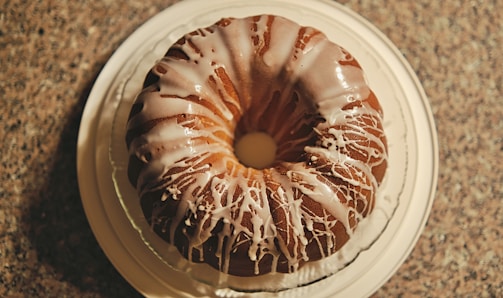 A round bundt cake with a drizzle of white icing on top, placed on a white plate. The cake is set on a textured countertop, creating a contrast with its smooth icing. The icing is unevenly spread, adding a sense of homemade charm.