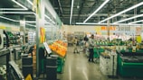 A supermarket interior with bright fluorescent lighting. Shoppers are browsing and pushing carts in an aisle filled with colorful produce on display. Signs indicate sections for meat and seafood, with other aisles visible in the background.