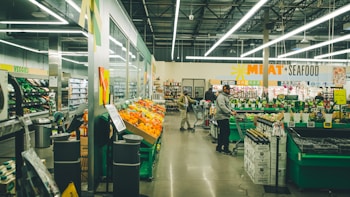 A supermarket interior with bright fluorescent lighting. Shoppers are browsing and pushing carts in an aisle filled with colorful produce on display. Signs indicate sections for meat and seafood, with other aisles visible in the background.