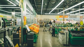 A supermarket interior with bright fluorescent lighting. Shoppers are browsing and pushing carts in an aisle filled with colorful produce on display. Signs indicate sections for meat and seafood, with other aisles visible in the background.