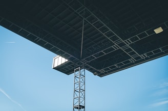 A large industrial structure with a metal framework is viewed from below against a clear blue sky. The structure includes a latticed metal column and a large flat surface, likely the underside of a roof or platform. A small light fixture and what appears to be a window or vent are visible. Additionally, a thin, white contrail is seen across the sky.