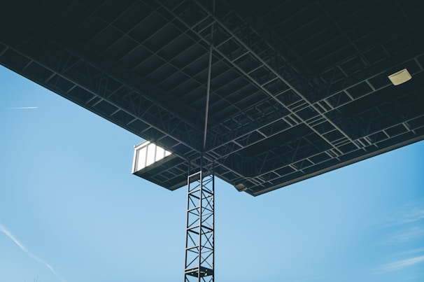 A large industrial structure with a metal framework is viewed from below against a clear blue sky. The structure includes a latticed metal column and a large flat surface, likely the underside of a roof or platform. A small light fixture and what appears to be a window or vent are visible. Additionally, a thin, white contrail is seen across the sky.