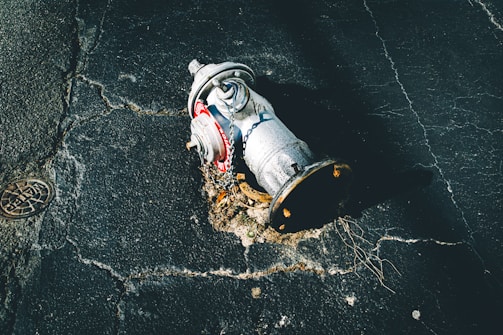 A damaged fire hydrant lies on its side on cracked asphalt. The metal surface is covered with rust and dirt, and a chain is attached to it. The pavement shows signs of wear and tear, with visible cracks and holes.