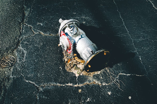 A damaged fire hydrant lies on its side on cracked asphalt. The metal surface is covered with rust and dirt, and a chain is attached to it. The pavement shows signs of wear and tear, with visible cracks and holes.