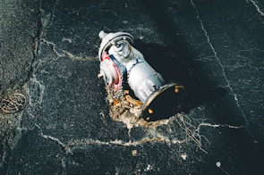 A damaged fire hydrant lies on its side on cracked asphalt. The metal surface is covered with rust and dirt, and a chain is attached to it. The pavement shows signs of wear and tear, with visible cracks and holes.