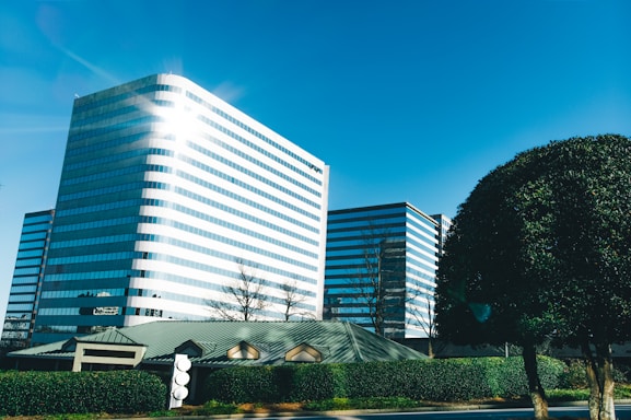 A modern office building with reflective glass windows stands tall against a clear blue sky. The sun creates a bright glare on the building's facade. In the foreground, neatly trimmed bushes and trees are visible alongside a smaller building with a green roof.