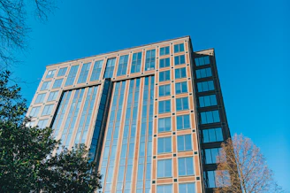 A modern office building with large glass windows reflecting the sky, surrounded by greenery.