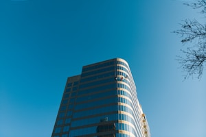 A tall, modern glass building with a curved facade stands against a clear blue sky. Two window cleaners on suspended platforms are visible midway up the building. The sunlight reflects off the windows, creating a bright and clean appearance. A bare tree branch is visible in the top right corner.