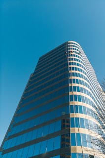 A modern high-rise building with reflective glass windows set against a clear blue sky. Two workers are visible, suspended near the top, likely engaged in cleaning or maintenance. The structure has a sleek, curving design and appears to be made of glass and steel.