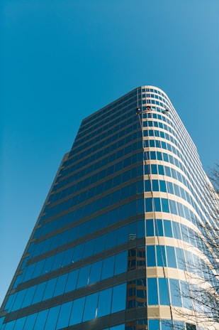 A modern high-rise building with reflective glass windows set against a clear blue sky. Two workers are visible, suspended near the top, likely engaged in cleaning or maintenance. The structure has a sleek, curving design and appears to be made of glass and steel.