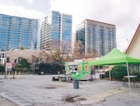 A cityscape featuring modern high-rise buildings in the background, with a food truck and a green canopy tent set up in an empty parking lot. Picnic tables and a large black vehicle are also present, suggesting a casual outdoor setting.