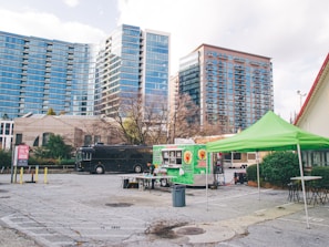A cityscape featuring modern high-rise buildings in the background, with a food truck and a green canopy tent set up in an empty parking lot. Picnic tables and a large black vehicle are also present, suggesting a casual outdoor setting.