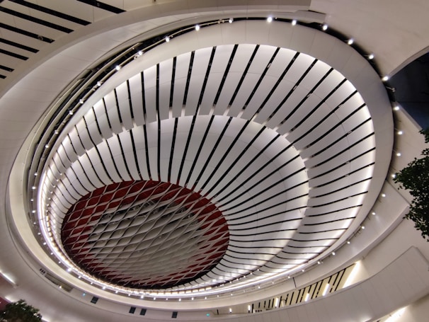 A modern architectural ceiling design featuring concentric circular patterns with alternating black and white stripes. The structure has a central circular opening with a red and white pattern of interlocking diamond shapes, surrounded by well-lit curved walls. The design creates a sense of depth and perspective in a spacious indoor area.