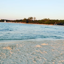 A serene beach scene with golden sand in the foreground, gentle waves lapping at the shore, and lush greenery lining the coast. The water is a clear blue, and the sky is a soft gradient of blue and orange, indicating either sunrise or sunset.