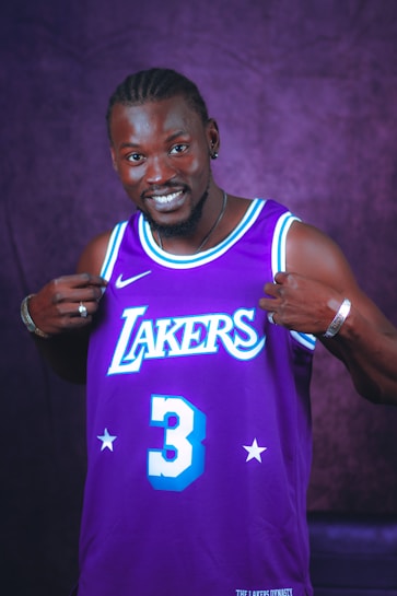 A person smiling while pointing at their purple sports jersey with 'Lakers' and the number 3 on it. The background is a matching purple color.