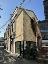A narrow street with a two-story building featuring a weathered exterior. The building has a sign that reads 'DELPHIC' and appears to have a small storefront with large glass windows. Two artificial trees are placed outside near the corner. The street is lined with wires and surrounded by other buildings, including a modern one in the background.