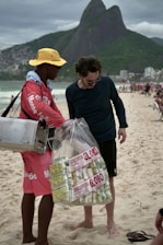 A street vendor is interacting with a customer on a beach. The vendor is wearing a red outfit and a yellow hat, carrying a metal container. He is holding a large bag filled with packaged snacks labeled 'Biscoitos Globo.' The customer is dressed in dark casual attire and is barefoot on the sand. In the background, there are mountains and a busy beachfront scene with other people.