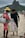 A street vendor is interacting with a customer on a beach. The vendor is wearing a red outfit and a yellow hat, carrying a metal container. He is holding a large bag filled with packaged snacks labeled 'Biscoitos Globo.' The customer is dressed in dark casual attire and is barefoot on the sand. In the background, there are mountains and a busy beachfront scene with other people.
