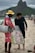 A street vendor is interacting with a customer on a beach. The vendor is wearing a red outfit and a yellow hat, carrying a metal container. He is holding a large bag filled with packaged snacks labeled 'Biscoitos Globo.' The customer is dressed in dark casual attire and is barefoot on the sand. In the background, there are mountains and a busy beachfront scene with other people.