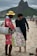 A street vendor is interacting with a customer on a beach. The vendor is wearing a red outfit and a yellow hat, carrying a metal container. He is holding a large bag filled with packaged snacks labeled 'Biscoitos Globo.' The customer is dressed in dark casual attire and is barefoot on the sand. In the background, there are mountains and a busy beachfront scene with other people.