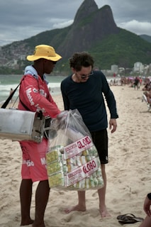 A street vendor is interacting with a customer on a beach. The vendor is wearing a red outfit and a yellow hat, carrying a metal container. He is holding a large bag filled with packaged snacks labeled 'Biscoitos Globo.' The customer is dressed in dark casual attire and is barefoot on the sand. In the background, there are mountains and a busy beachfront scene with other people.