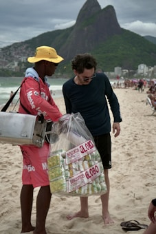 A street vendor is interacting with a customer on a beach. The vendor is wearing a red outfit and a yellow hat, carrying a metal container. He is holding a large bag filled with packaged snacks labeled 'Biscoitos Globo.' The customer is dressed in dark casual attire and is barefoot on the sand. In the background, there are mountains and a busy beachfront scene with other people.