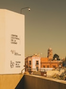 An urban scene with a tall white building on the left displaying the names 'Centro Cultural de la Ciudad', 'Cineteca Rosalío Solano', and 'Teatro de la Ciudad'. In the background, there is a red-orange building and a church with a dome and cross. A security camera is mounted on the white building.
