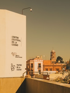 An urban scene with a tall white building on the left displaying the names 'Centro Cultural de la Ciudad', 'Cineteca Rosalío Solano', and 'Teatro de la Ciudad'. In the background, there is a red-orange building and a church with a dome and cross. A security camera is mounted on the white building.