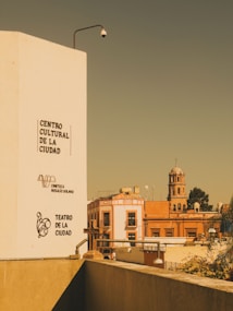 An urban scene with a tall white building on the left displaying the names 'Centro Cultural de la Ciudad', 'Cineteca Rosal&iacute;o Solano', and 'Teatro de la Ciudad'. In the background, there is a red-orange building and a church with a dome and cross. A security camera is mounted on the white building.