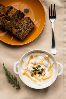 A cozy breakfast setting featuring a plate with slices of banana bread and a bowl of yogurt topped with slices of fruit and pumpkin seeds. A fork is placed beside the bowl on a beige tablecloth, accompanied by a small sprig of greenery.