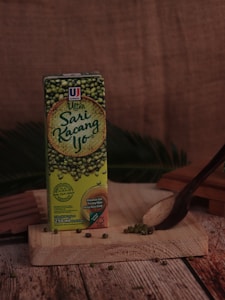 A carton of Ultra Sari Kacang Ijo, a mung bean drink, is placed on a wooden board. Green mung beans are scattered on the board with a dark wooden spoon nearby. The background is made of burlap fabric, enhancing the rustic feel.