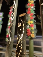 Decorative bamboo stalks with intricate patterns cut into them are displayed. They are illuminated from within, creating a warm glow. Some of the surrounding stalks are adorned with colorful flowers that feature red, yellow, and white blooms.