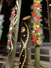 Decorative bamboo stalks with intricate patterns cut into them are displayed. They are illuminated from within, creating a warm glow. Some of the surrounding stalks are adorned with colorful flowers that feature red, yellow, and white blooms.