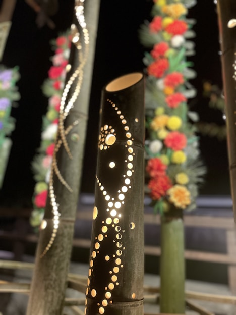 Decorative bamboo stalks with intricate patterns cut into them are displayed. They are illuminated from within, creating a warm glow. Some of the surrounding stalks are adorned with colorful flowers that feature red, yellow, and white blooms.
