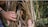 A close-up view of a tree trunk surrounded by dried palm fronds, with two hands gently pulling the fronds apart to reveal the tree. A large insect, possibly a cockroach, is visible nestled in the crevice of the trunk. The background includes blurred greenery.