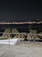 A nighttime scene featuring fireworks equipment arranged on platforms over water. The background reveals a cityscape with numerous lights from buildings reflecting on the water. Two people stand nearby the equipment, possibly preparing for an event.