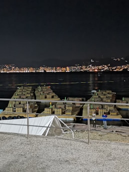 A nighttime scene featuring fireworks equipment arranged on platforms over water. The background reveals a cityscape with numerous lights from buildings reflecting on the water. Two people stand nearby the equipment, possibly preparing for an event.