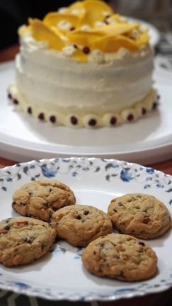 A beautifully decorated cake with white icing and yellow flower-like decorations is placed on a white platter, surrounded by small dark red edible accents around the base. In front of it, there is a decorative plate with a floral pattern, holding six golden-brown cookies with chocolate chips.