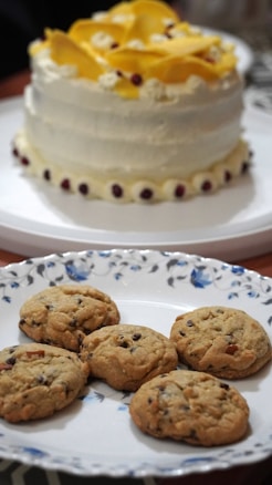 A beautifully decorated cake with white icing and yellow flower-like decorations is placed on a white platter, surrounded by small dark red edible accents around the base. In front of it, there is a decorative plate with a floral pattern, holding six golden-brown cookies with chocolate chips.