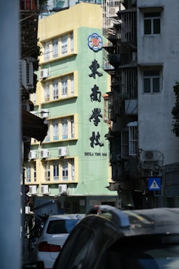 A city street with parked cars and a prominent building in the background. The building has a tall, green façade adorned with large Chinese characters and a logo at the top. The structure features multiple windows equipped with air conditioning units. There is visible signage in multiple languages including Portuguese. The lighting suggests it is daytime.