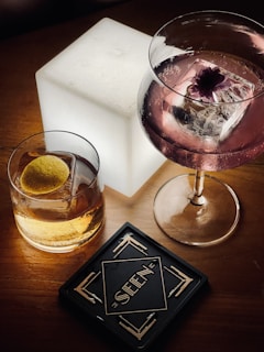 A wooden table features two elegant alcoholic drinks, one in a short glass with a lemon slice and another in a stemmed glass with a pink liquid and a decorative flower atop a large ice cube. A glowing white cube light adds ambiance, and a cocktail coaster with Art Deco design displays the word 'SEEN'.