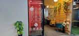 An office space features a wall with a large digital screen displaying an oversized phone dial pad. To the right, there is a cozy seating area with a teal chair and wooden elements adorned with decorative plants. A potted plant is positioned against the opposite wall, which is plain and light-colored.