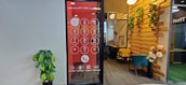 An office space features a wall with a large digital screen displaying an oversized phone dial pad. To the right, there is a cozy seating area with a teal chair and wooden elements adorned with decorative plants. A potted plant is positioned against the opposite wall, which is plain and light-colored.