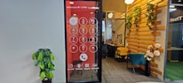 An office space features a wall with a large digital screen displaying an oversized phone dial pad. To the right, there is a cozy seating area with a teal chair and wooden elements adorned with decorative plants. A potted plant is positioned against the opposite wall, which is plain and light-colored.