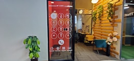 An office space features a wall with a large digital screen displaying an oversized phone dial pad. To the right, there is a cozy seating area with a teal chair and wooden elements adorned with decorative plants. A potted plant is positioned against the opposite wall, which is plain and light-colored.