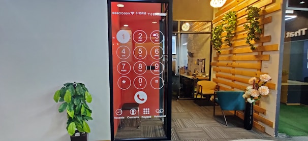 An office space features a wall with a large digital screen displaying an oversized phone dial pad. To the right, there is a cozy seating area with a teal chair and wooden elements adorned with decorative plants. A potted plant is positioned against the opposite wall, which is plain and light-colored.