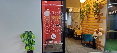 An office space features a wall with a large digital screen displaying an oversized phone dial pad. To the right, there is a cozy seating area with a teal chair and wooden elements adorned with decorative plants. A potted plant is positioned against the opposite wall, which is plain and light-colored.