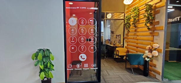 An office space features a wall with a large digital screen displaying an oversized phone dial pad. To the right, there is a cozy seating area with a teal chair and wooden elements adorned with decorative plants. A potted plant is positioned against the opposite wall, which is plain and light-colored.