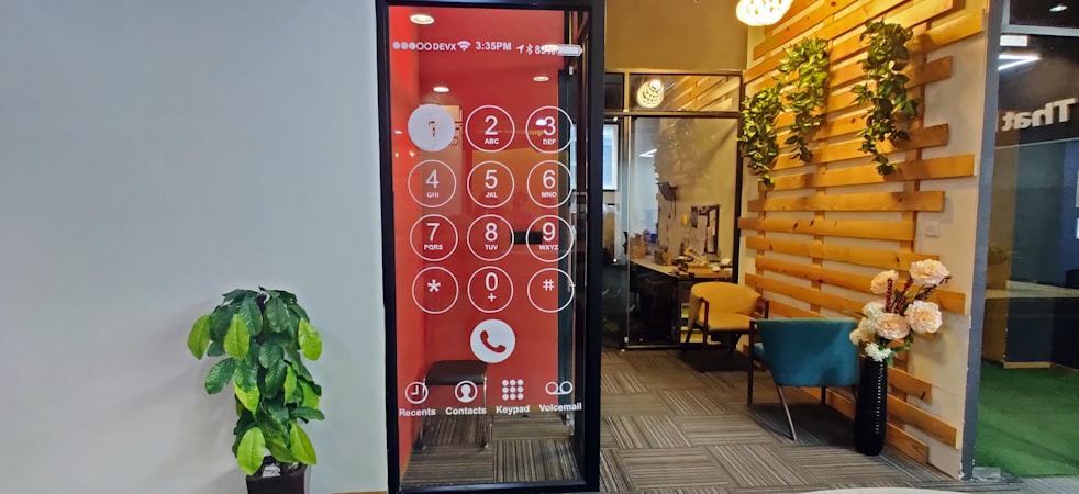 An office space features a wall with a large digital screen displaying an oversized phone dial pad. To the right, there is a cozy seating area with a teal chair and wooden elements adorned with decorative plants. A potted plant is positioned against the opposite wall, which is plain and light-colored.