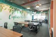 An open office workspace featuring several desks with ergonomic chairs. The walls are decorated with a nature-themed mural and the phrase 'Capture the Nature' is prominently displayed. Overhead, modern light fixtures and exposed ductwork add an industrial touch. Natural light filters in through large windows, enhancing the workspace.